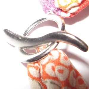 May include: A silver-toned metal toggle clasp featuring a circular ring and a curved bar component. The clasp is attached to fabric elements with a vibrant orange and white patterned design, alongside a section of purple and orange fabric.