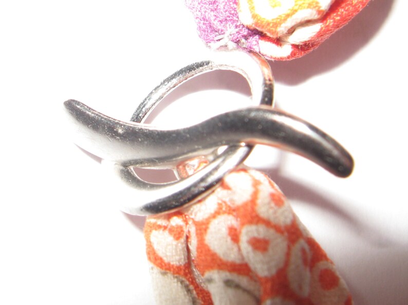 May include: A silver-toned metal toggle clasp featuring a circular ring and a curved bar component. The clasp is attached to fabric elements with a vibrant orange and white patterned design, alongside a section of purple and orange fabric.