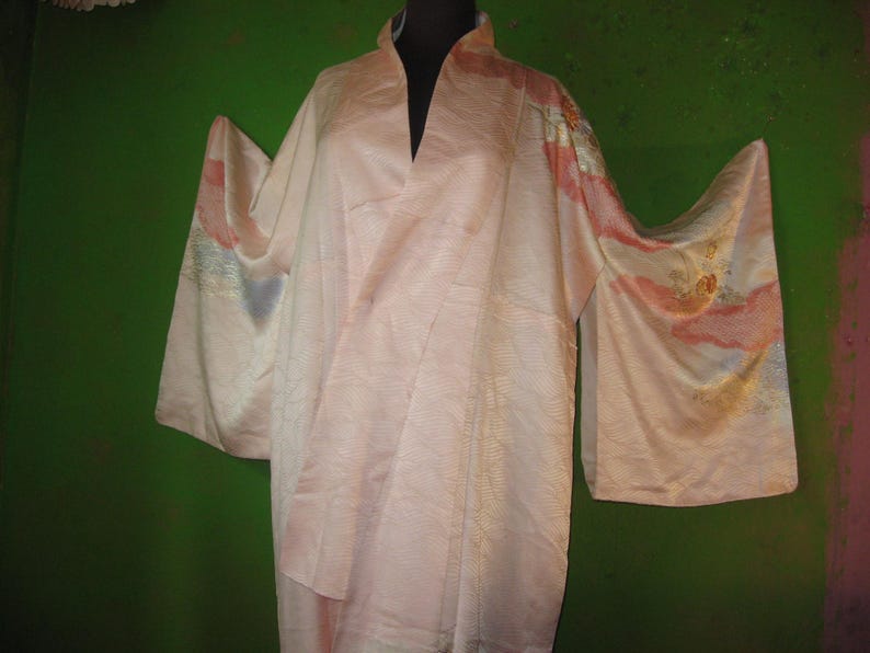 May include: A pale pink kimono with a V-neck and long sleeves. The kimono has a subtle floral pattern in shades of pink, blue, and gold.