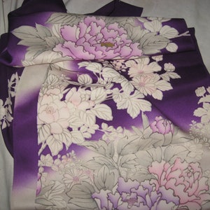 May include: A folded purple kimono with a floral pattern. The design features large, light purple and pink peony flowers with white and gray leaves. The fabric has a gradient effect, transitioning from a deep purple to a lighter shade.