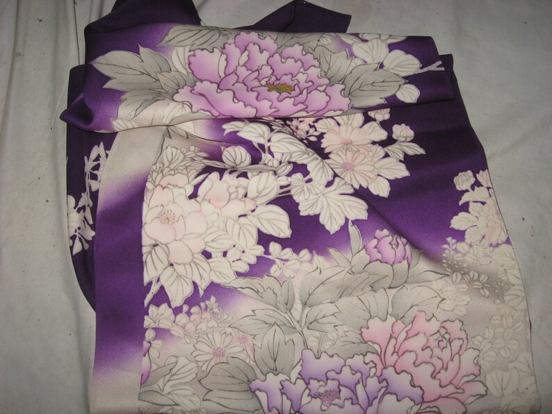May include: A folded purple kimono with a floral pattern. The design features large, light purple and pink peony flowers with white and gray leaves. The fabric has a gradient effect, transitioning from a deep purple to a lighter shade.