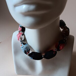 May include: A necklace featuring cylindrical fabric beads with various patterns including black, red, and blue floral and geometric designs, separated by small silver-toned metal spacer beads, arranged in a continuous circular strand around the neck.