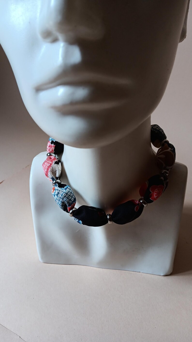 May include: A necklace featuring cylindrical fabric beads with various patterns including black, red, and blue floral and geometric designs, separated by small silver-toned metal spacer beads, arranged in a continuous circular strand around the neck.