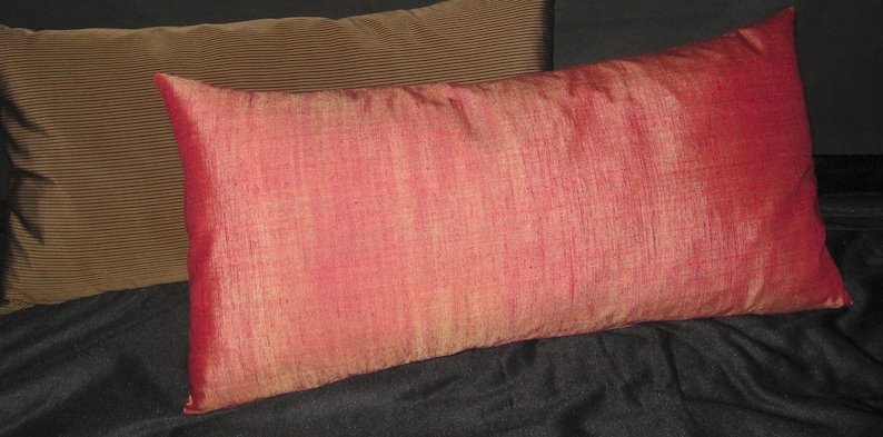 May include: Two rectangular throw pillows. The front pillow is a vibrant red with a textured appearance. The second pillow is a muted brown with a ribbed design. Both pillows are on a dark surface.