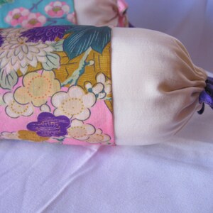 May include: A long, cylindrical pillow with a floral print fabric cover. The pillow has a cream-colored fabric lining and a purple ribbon tied around the end.