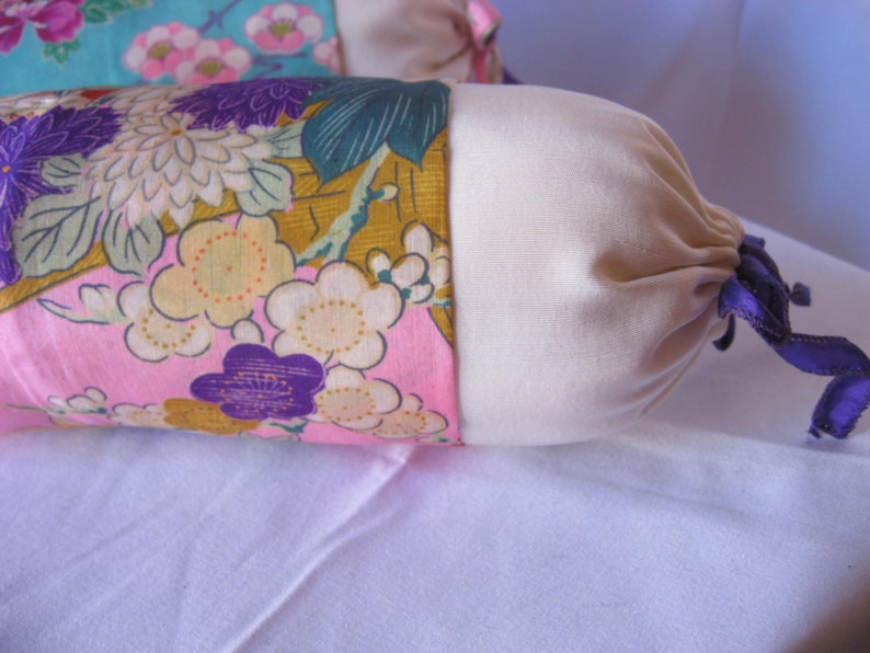 May include: A long, cylindrical pillow with a floral print fabric cover. The pillow has a cream-colored fabric lining and a purple ribbon tied around the end.