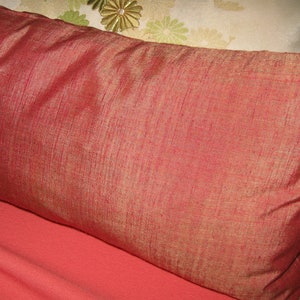 May include: A rectangular, reddish-orange pillow with a textured, silk-like appearance. The pillow is resting on a coral-colored surface. Another pillow with a floral design is partially visible in the background.
