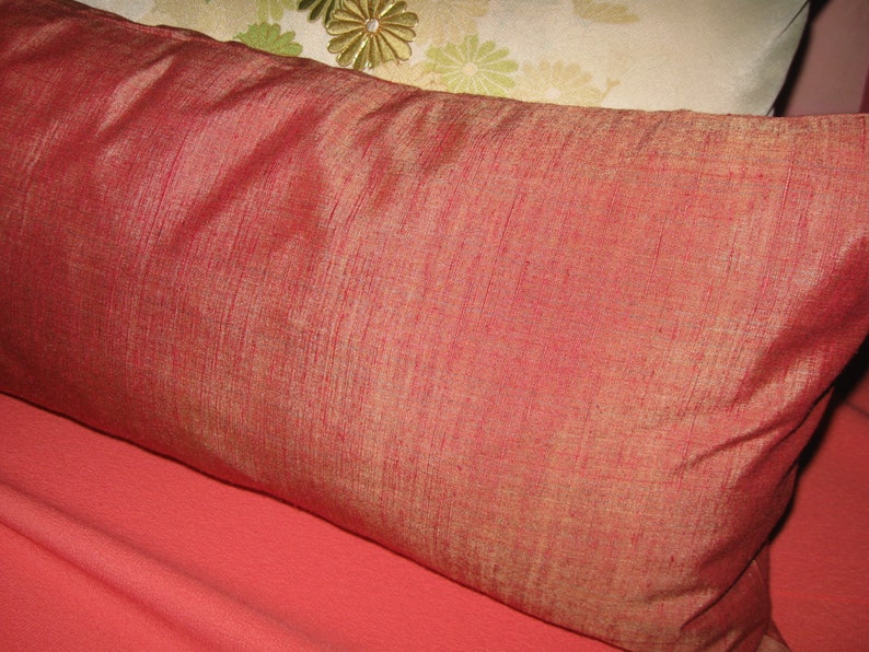 May include: A rectangular, reddish-orange pillow with a textured, silk-like appearance. The pillow is resting on a coral-colored surface. Another pillow with a floral design is partially visible in the background.