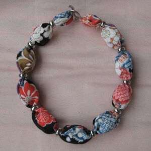 May include: A necklace featuring a series of fabric-covered beads in various floral and geometric patterns, including red, blue, white, and black colors. The beads are separated by small, round, silver-toned metal spacers and secured with a metal clasp.