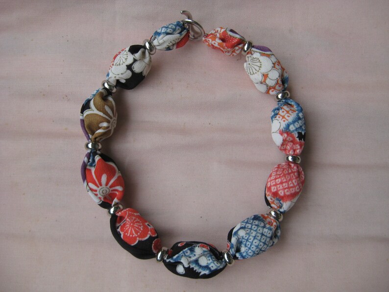 May include: A necklace featuring a series of fabric-covered beads in various floral and geometric patterns, including red, blue, white, and black colors. The beads are separated by small, round, silver-toned metal spacers and secured with a metal clasp.