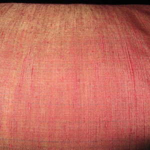 May include: Close-up of a textured, reddish-pink fabric. The material has a subtle sheen and a woven pattern with vertical lines. The color shifts slightly to a golden hue in areas, creating a visual depth. The fabric appears to be a decorative textile.