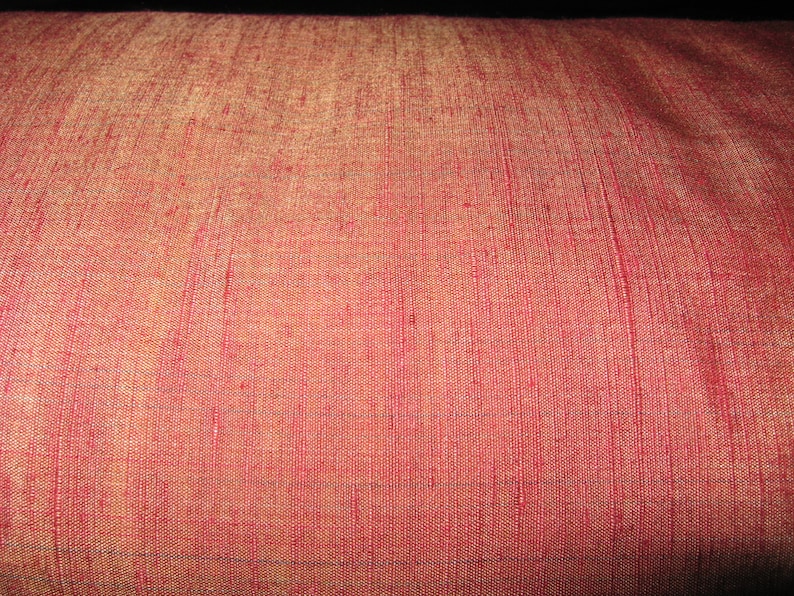 May include: Close-up of a textured, reddish-pink fabric. The material has a subtle sheen and a woven pattern with vertical lines. The color shifts slightly to a golden hue in areas, creating a visual depth. The fabric appears to be a decorative textile.