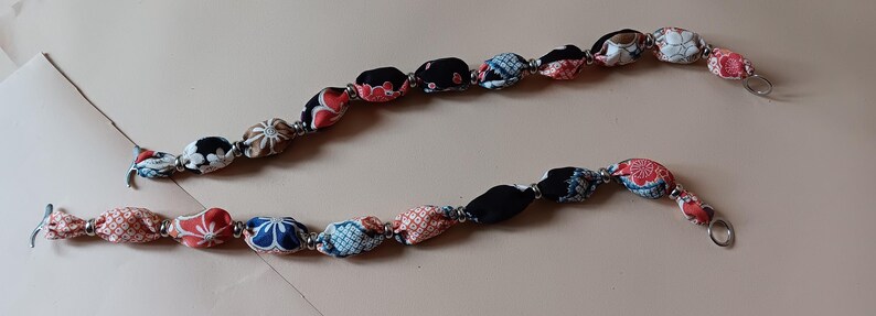 May include: Two decorative bag straps featuring a series of fabric-covered beads in black, red, white, and blue with floral and geometric patterns. Each bead is separated by small silver-toned metal spacers, and the ends feature silver metal rings.