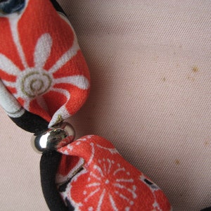 May include: A fabric necklace featuring segments of orange, black, and white floral patterned textile separated by smooth, polished silver-toned metal beads. The soft fabric sections are knotted or gathered through the metallic spacers.