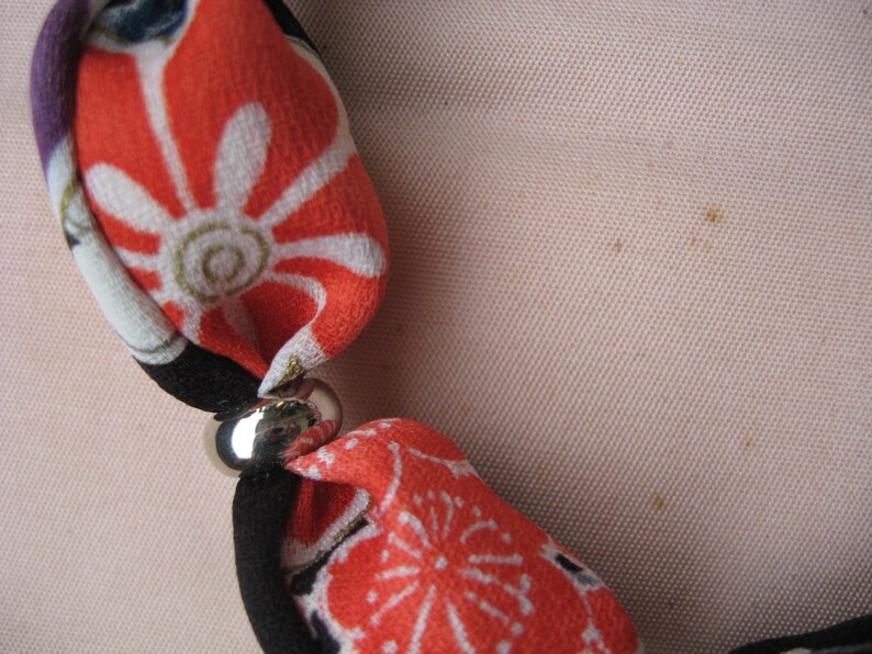 May include: A fabric necklace featuring segments of orange, black, and white floral patterned textile separated by smooth, polished silver-toned metal beads. The soft fabric sections are knotted or gathered through the metallic spacers.