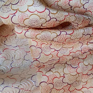 Könnte beinhalten: Cremefarbener Stoff mit einem sich wiederholenden Blumenmuster in Rot, Gold und Lila. Der Stoff hat ein strukturiertes Aussehen und ist drapiert, wodurch das komplizierte Design und die Falten sichtbar werden.