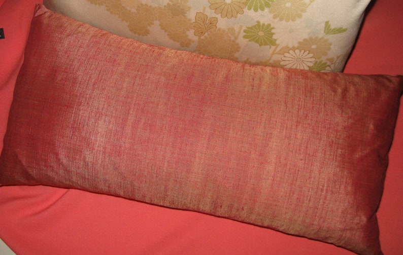 May include: A rectangular, reddish-orange pillow with a textured surface. The pillow is placed on a coral-colored surface, with a floral patterned pillow in the background. The pillow is likely for home decor.