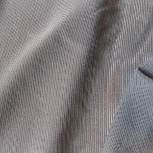 K&ouml;nnte beinhalten: Ein dunkel anthrazitfarbener Stoff mit einer feinen vertikalen Rippenstruktur. Das Material weist weiche Falten und Drapierungen auf, die das gleichm&auml;&szlig;ige lineare Muster auf der Oberfl&auml;che hervorheben. Das Textil hat eine matte Oberfl&auml;che mit dezenten Lichtreflexen.