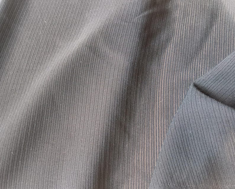 K&ouml;nnte beinhalten: Ein dunkel anthrazitfarbener Stoff mit einer feinen vertikalen Rippenstruktur. Das Material weist weiche Falten und Drapierungen auf, die das gleichm&auml;&szlig;ige lineare Muster auf der Oberfl&auml;che hervorheben. Das Textil hat eine matte Oberfl&auml;che mit dezenten Lichtreflexen.