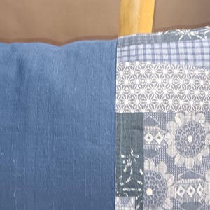 Puede incluir: Un cojín rectangular con tela de lino azul en un lado y un diseño de patchwork en el otro. El patchwork presenta patrones florales y geométricos en tonos azules y grises. Un elemento decorativo para el hogar.