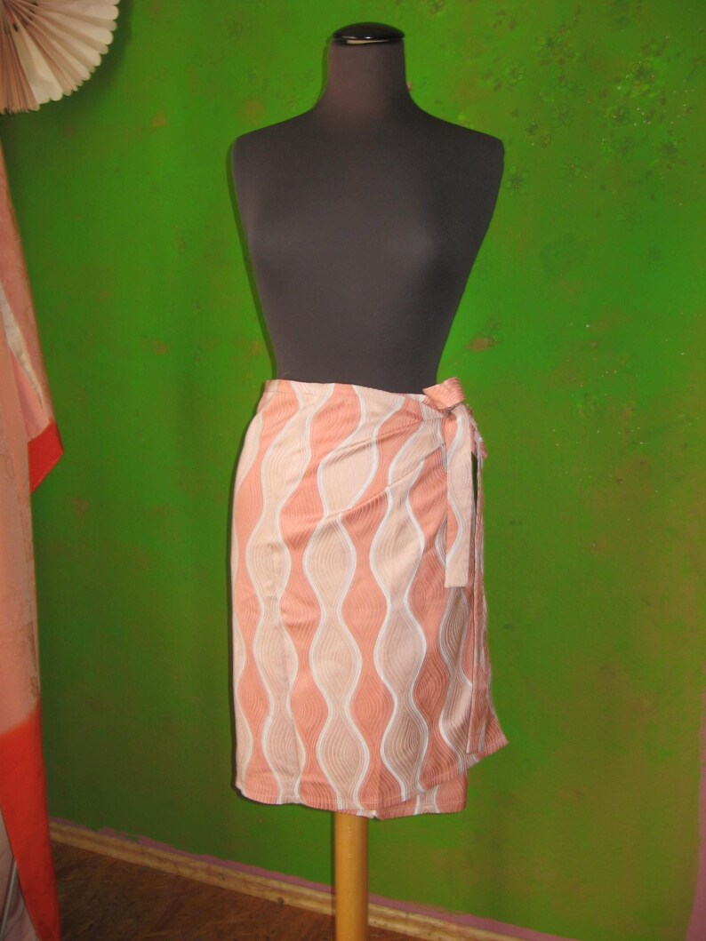 May include: A wrap skirt featuring a peach and light pink wavy vertical pattern with white outlines. The skirt has a side tie closure and falls to knee length, displaying a consistent repeating graphic design across the fabric surface.