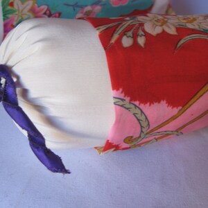 May include: A white cylindrical pillow with a purple ribbon tied around it. The pillow is covered in a red fabric with a floral pattern in pink, white, and gold.