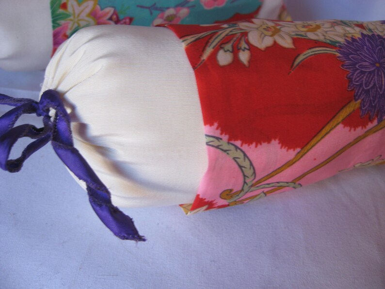 May include: A white cylindrical pillow with a purple ribbon tied around it. The pillow is covered in a red fabric with a floral pattern in pink, white, and gold.