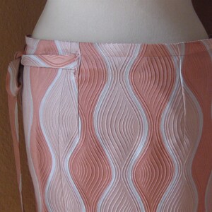 May include: A wrap skirt featuring a textured, wavy vertical pattern in shades of peach, light pink, and white. The garment has a side tie closure at the waistband and a structured, embossed fabric surface that creates a repeating undulating design.
