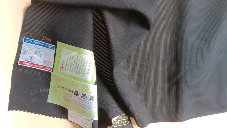 K&ouml;nnte beinhalten: Ein dunkel anthrazitfarbener Stoff mit einer feinen, vertikal gerippten Textur. Am Stoff sind mehrere rechteckige Etiketten mit japanischem Text befestigt, darunter ein blau-wei&szlig;es Etikett mit Illustrationen und ein hellgr&uuml;nes Zertifikat.