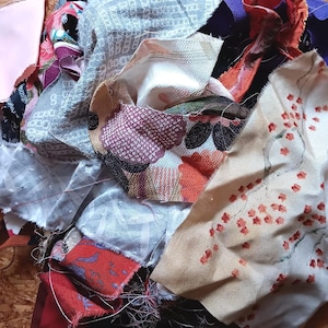 Könnte beinhalten: Eine Sammlung verschiedener Stoffreste mit unterschiedlichen Mustern und Farben, darunter ein hellbeiges Stück mit rotem Blütenzweig-Druck, ein dunkelvioletter Streifen, ein graues geometrisches Musterstück und verschiedene andere strukturierte Textilreste.