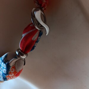 May include: A necklace featuring fabric-covered beads in shades of red, blue, and white with a floral pattern. The beads are separated by silver-toned metal rings and secured with a silver-toned metal hook clasp, creating a continuous chain design.