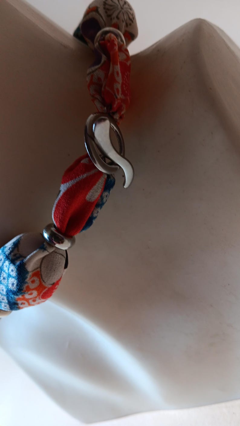 May include: A necklace featuring fabric-covered beads in shades of red, blue, and white with a floral pattern. The beads are separated by silver-toned metal rings and secured with a silver-toned metal hook clasp, creating a continuous chain design.