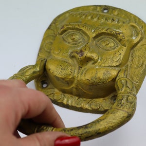 Puede incluir: Un llamador de puerta de hierro fundido de color dorado con una cara detallada y un asa de anillo. El llamador tiene la forma de una cabeza de león con una superficie texturizada.