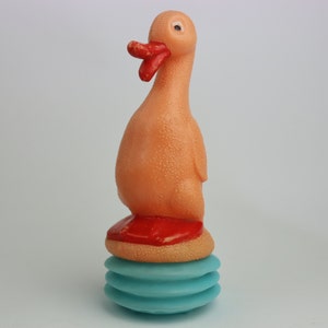 May include: A pink rubber duck toy with a red beak and a blue and white base. The duck is sitting on a blue and white base with a red ring around the top.