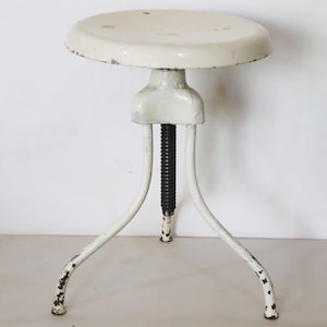 Pode incluir: Banqueta industrial vintage creme com assento redondo e três pernas curvas. A banqueta apresenta uma haste roscada preta ajustável e mostra sinais de desgaste. A estrutura metálica tem um acabamento envelhecido.