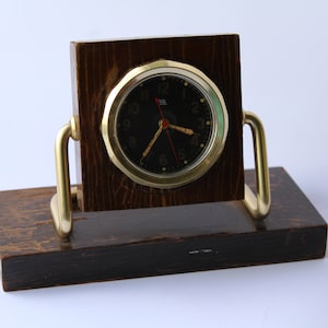 Pode incluir: Um relógio vintage com um mostrador preto e ponteiros dourados, montado em um suporte de madeira escura com detalhes em latão.