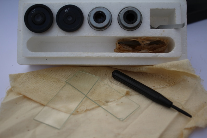 May include: A white foam box containing four microscope lenses, two glass slides, and a black tool. The lenses are labelled "10x" and "15x".