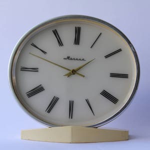May include: A silver and white analogue clock with black Roman numeral hour markers and a gold-coloured hour and minute hand. The clock face reads "Маяк" in Cyrillic script.