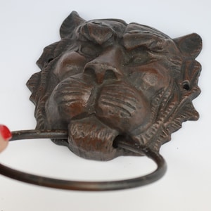 Puede incluir: Un toallero de cabeza de tigre de hierro fundido con un acabado marrón oscuro. El anillo es de metal y tiene un diseño sencillo.