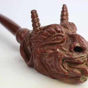 Puede incluir: Una pipa de madera con una cara grotesca tallada. La pipa es marrón y tiene una boquilla larga y curva. La cara tiene una boca ancha, una nariz larga y dos cuernos.