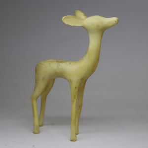 May include: A pale yellow deer figurine with a slender neck and long legs. The deer has small, upright ears and a slightly raised head, giving it a graceful appearance. The figurine is made of a solid material and stands on four legs.