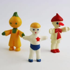 May include: Three vintage plastic figurines. A yellow and green figure with a rounded head, a white figure with a red star on its chest, and a white figure with a red hat and a red bow.