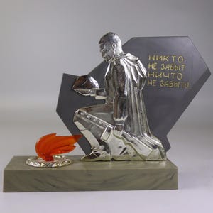 May include: A silver sculpture of a kneeling soldier holding a helmet, with a flame and text on a dark gray background. The text is in gold and reads "Никто не забыт, ничто не забыто."