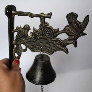 Puede incluir: Campana de pared de hierro fundido negro con un diseño de pájaro y flores. La campana tiene una cadena unida a la parte inferior.