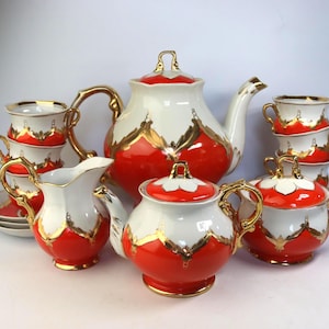 Könnte beinhalten: Ein Vintage-Teeservice in Rot, Weiß und Gold. Das Set umfasst eine Teekanne, eine Zuckerdose, einen Sahnekännchen, Tassen und Untertassen. Die Teile haben goldfarbene Verzierungen und dekorative Details.