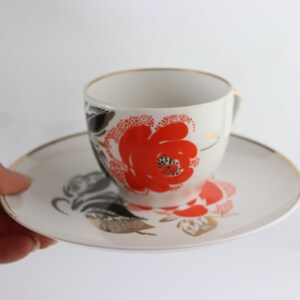 May include: A white teacup and saucer set with a floral design. The cup and saucer feature a red and black floral pattern with gold accents.