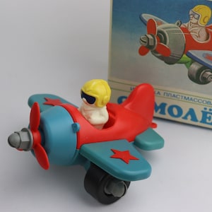 May include: A red and blue toy airplane with a yellow pilot figure. The airplane has a propeller and a red star on the wing. The toy is made of plastic and has a simple design.