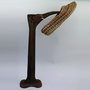 May include: A vintage metal shoe stretcher with a woven straw foot form. The stretcher has a black metal base and a long metal arm with a hinged foot form.