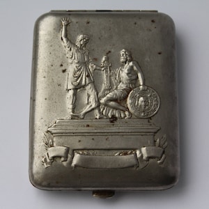 May include: A silver metal cigarette case with an embossed design of two figures standing on a pedestal. The figures are holding a torch and a shield with a profile of a person on it.