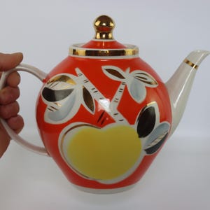 Könnte beinhalten: Eine lebendige, vintage Teekanne mit rotem Grund, verziert mit goldenen, weißen und schwarzen floralen Designs. Die Teekanne hat einen weißen Griff und Ausguss sowie einen goldfarbenen Deckelknauf. Ein gelbes herzförmiges Design ist vorhanden.